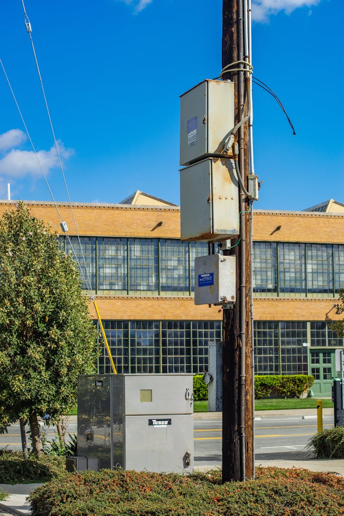Utility pole with metal boxes in front of a brick building on a sunny day.